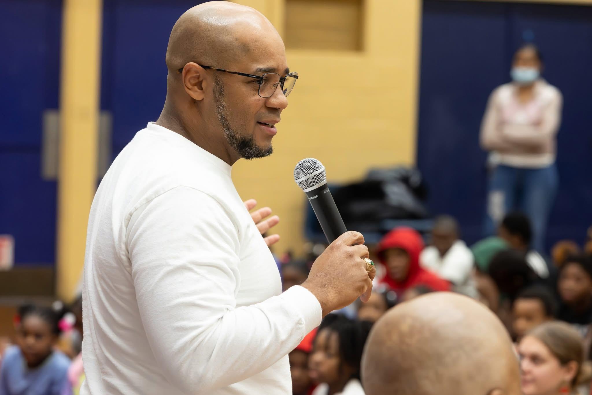 Mike Brown speaking into a microphone to a room full of seated students in a school gym